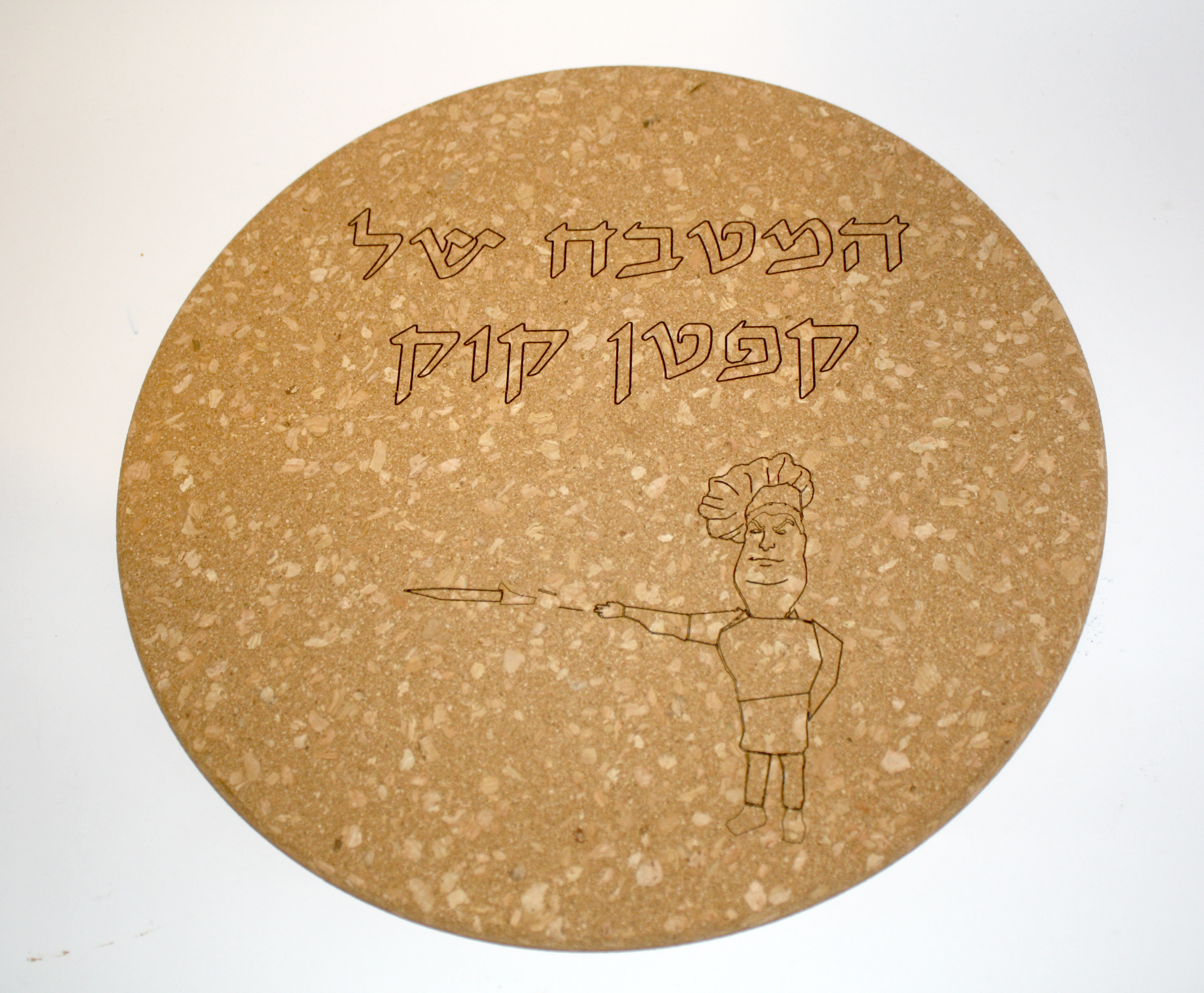 משטח שעם עגול לסירים חמים עם חריטת לייזר בשורות ישרות. בחלק העליון כתוב "המטבח של קפטן קוק" בפונט סופר סתם עם קו מתאר כפול, ובתחתית איור חרוט של דמות שף קומי מצביע עם סכין.